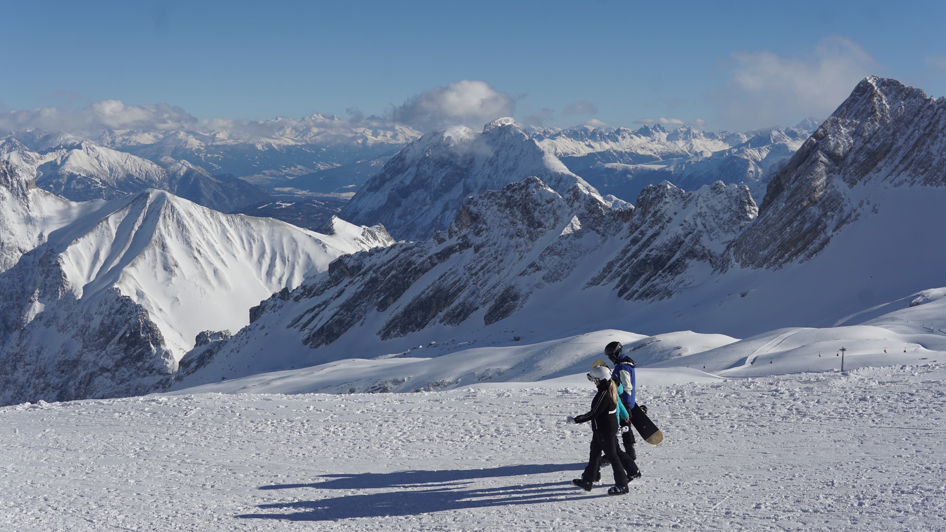 Snowboarders walking across a snow-covered alpine summit with dramatic mountain peaks in the background
