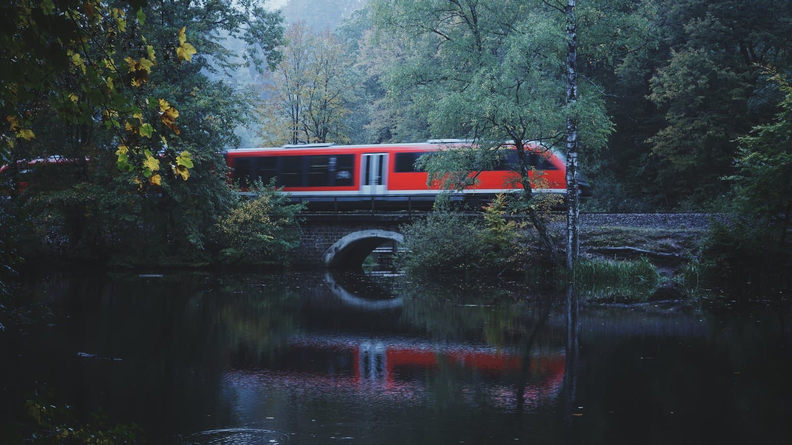 Red train crossing a stone bridge reflected in still water surrounded by misty forest