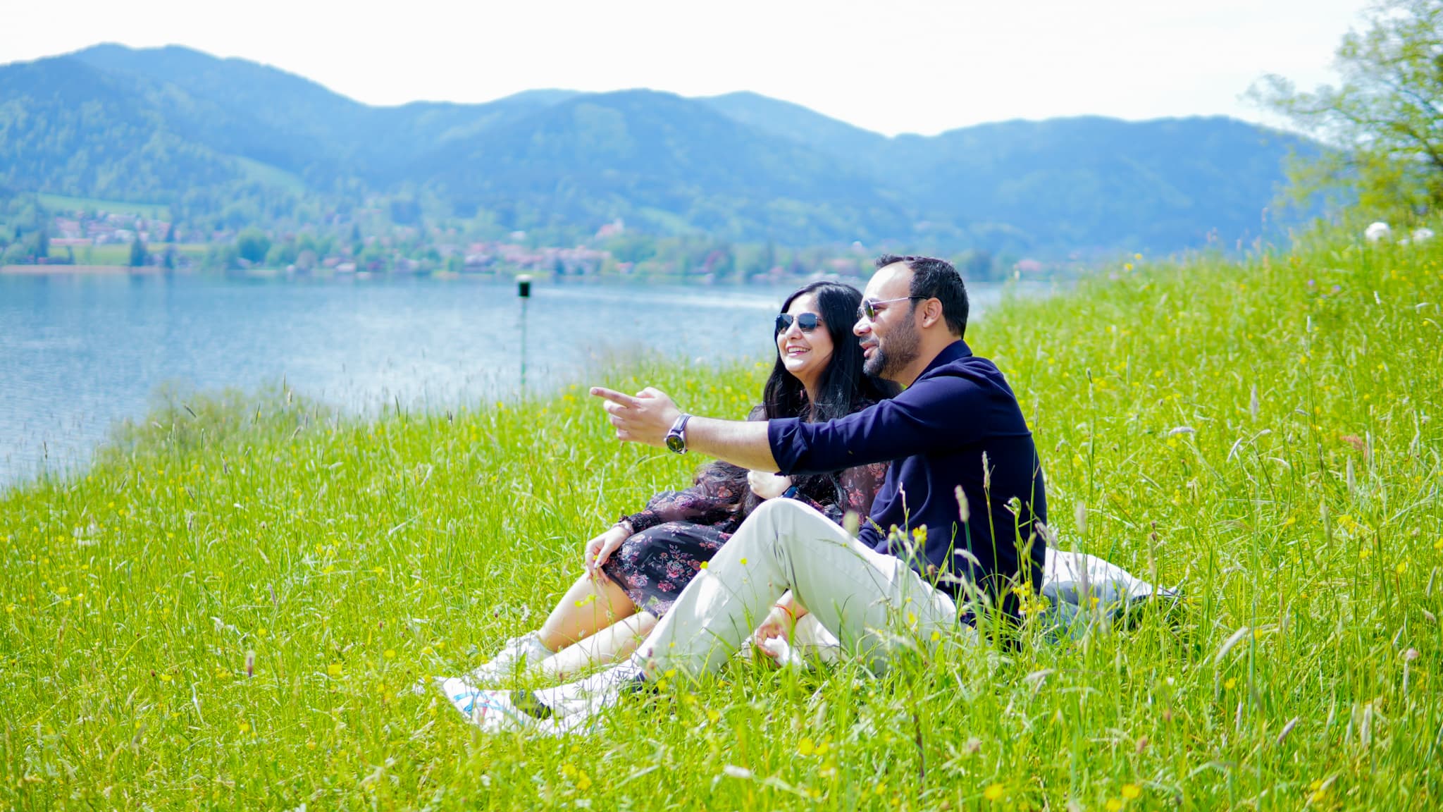 Couple sitting together on lush green grass by an alpine lake, pointing toward the distant mountains on a sunny day