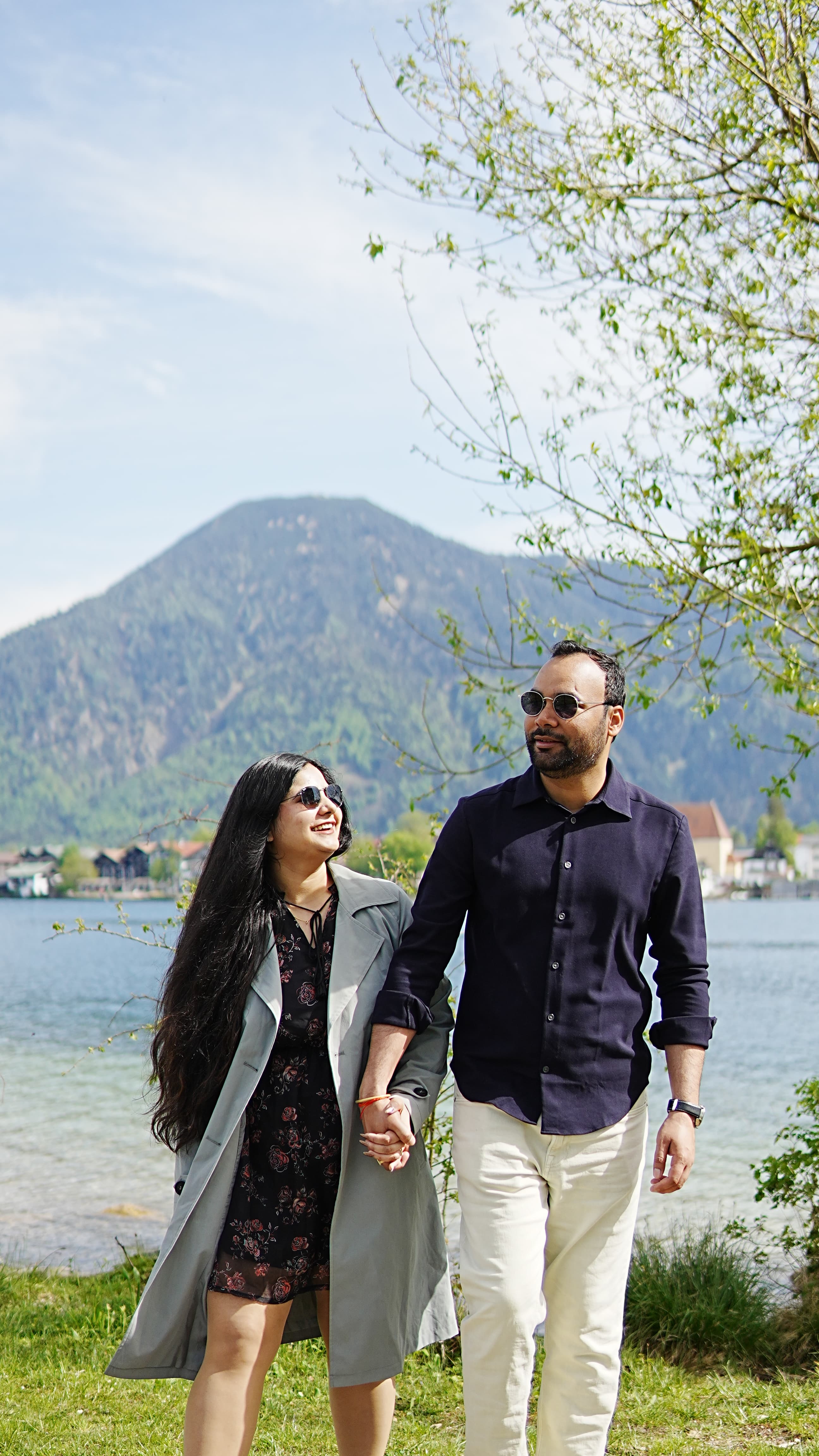 Couple walking hand in hand along a lakeside path with mountains and blue sky behind them