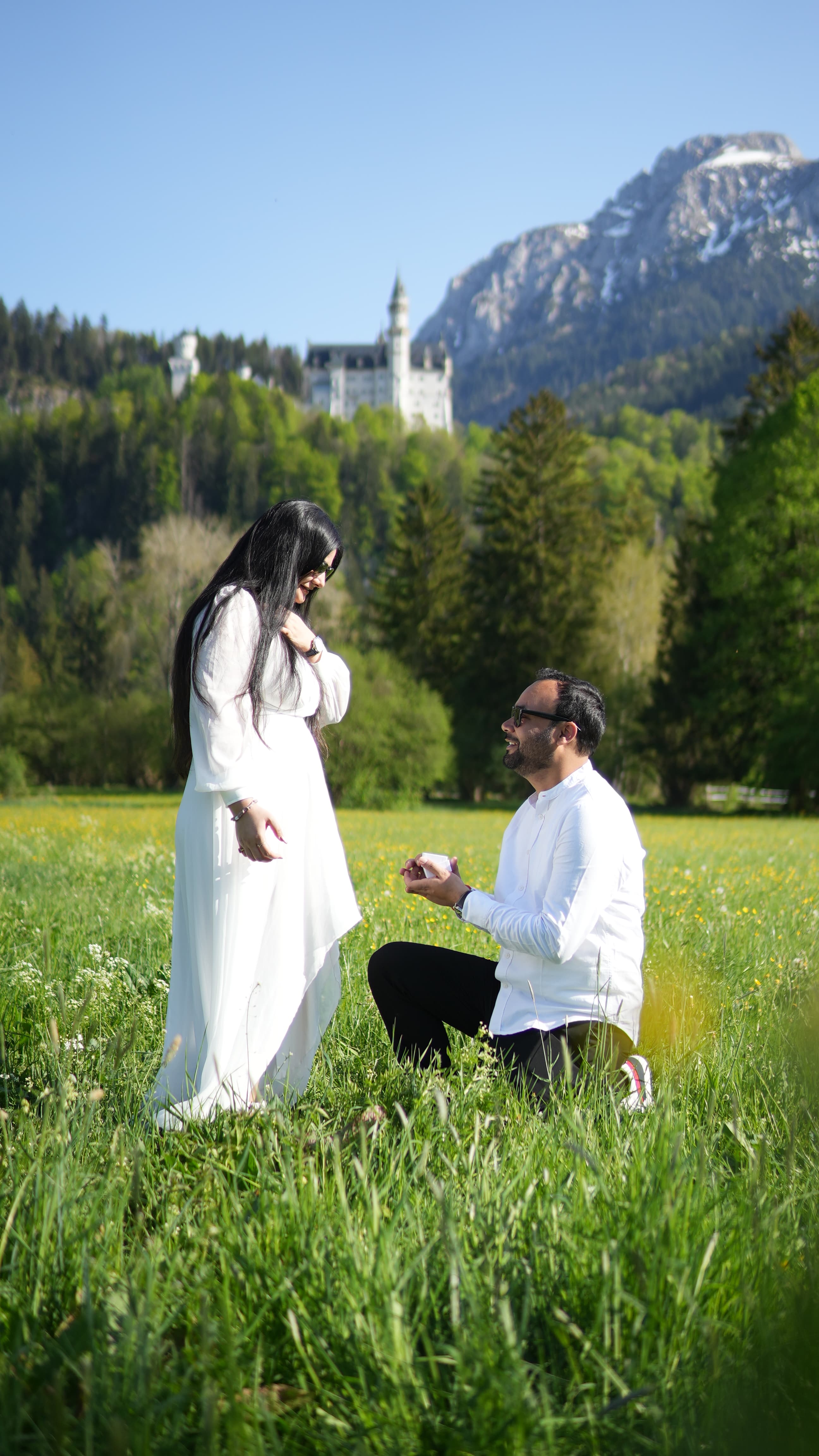 Couple in white walking through a sunlit green meadow with snow-capped mountains and a lake in the background