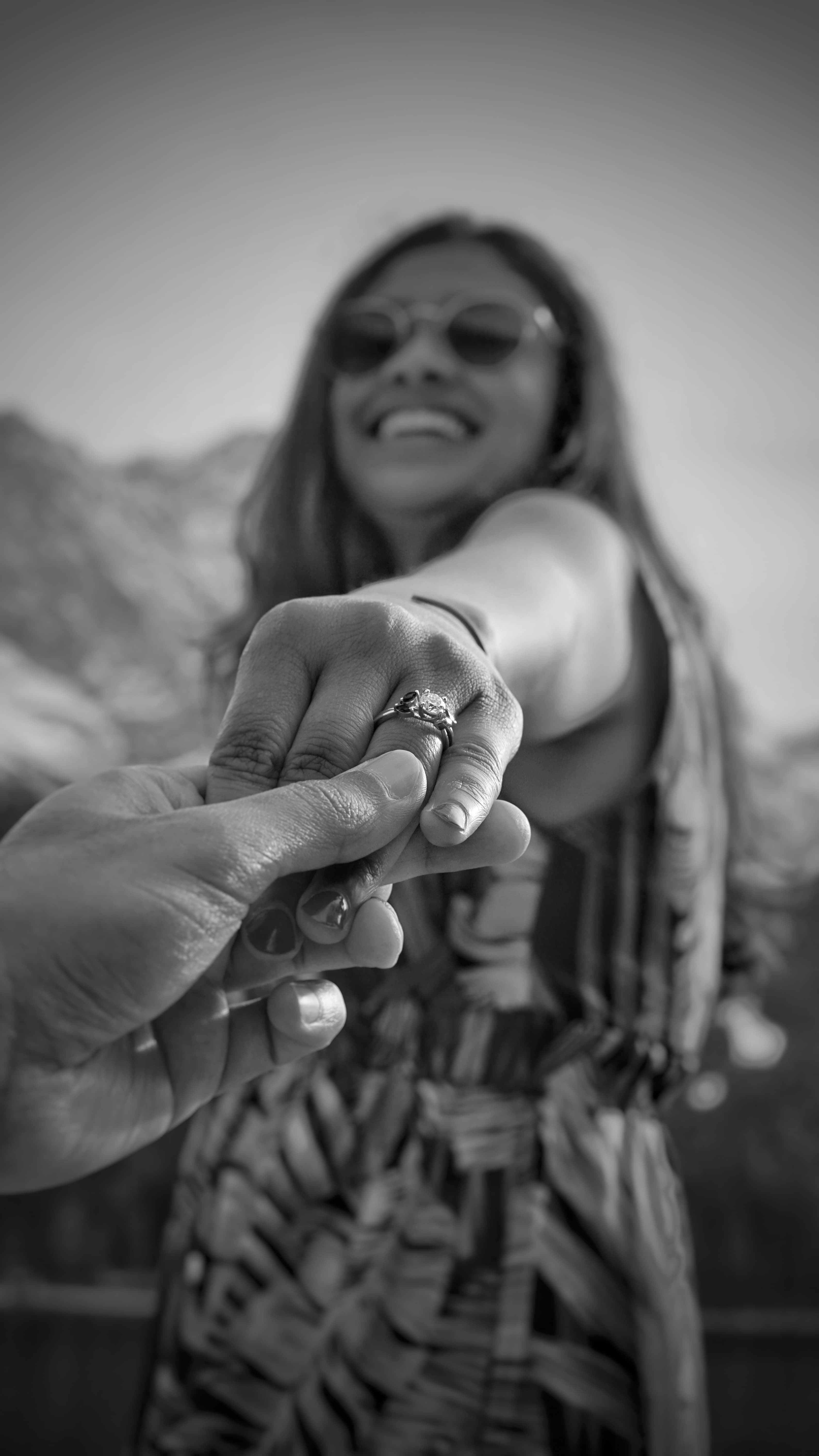 Black and white close-up of a smiling woman reaching toward the camera showing her new engagement ring with mountains in the background