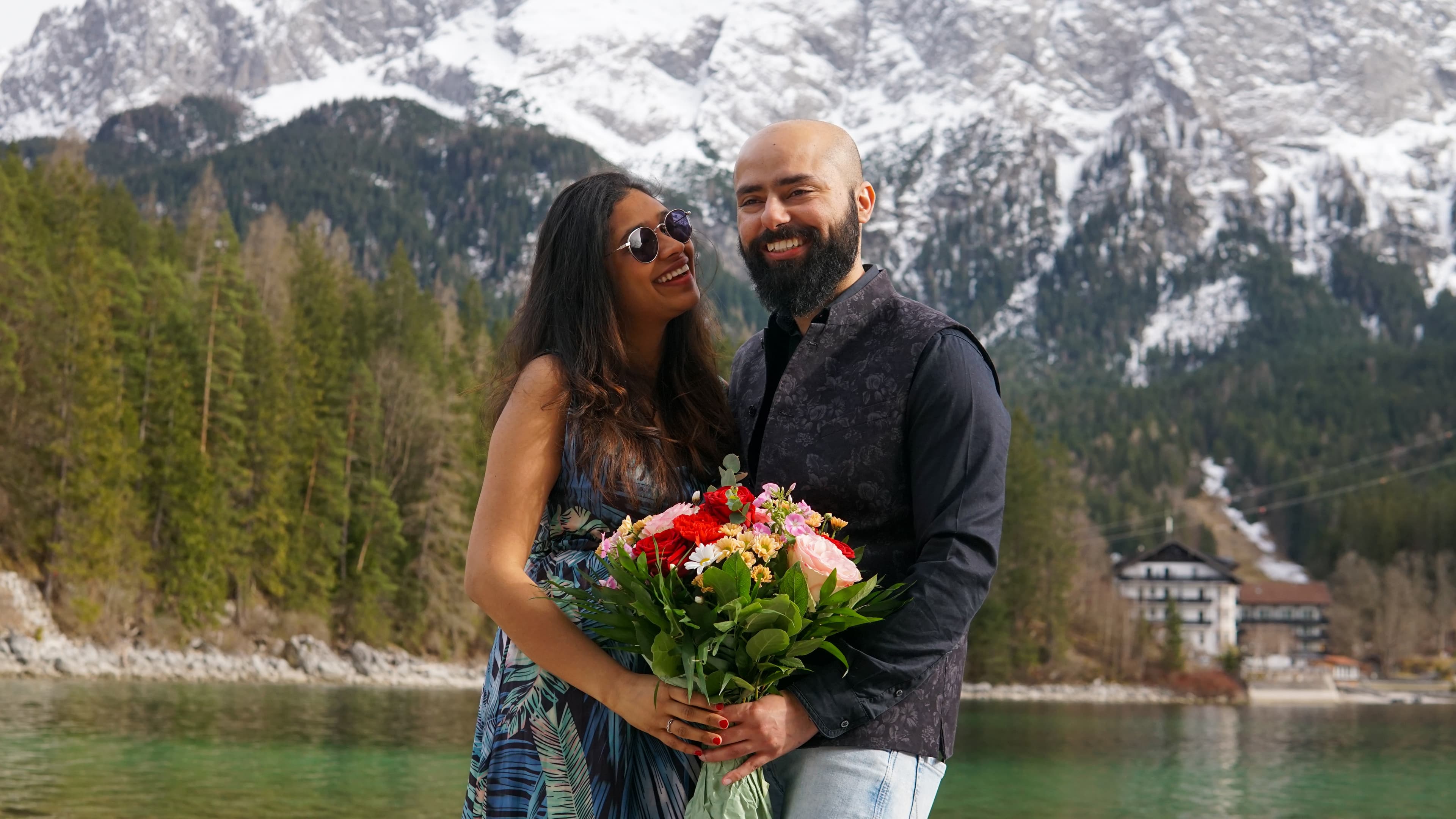 Newly engaged couple beaming with a colorful bouquet of flowers by a turquoise alpine lake beneath snow-covered mountains