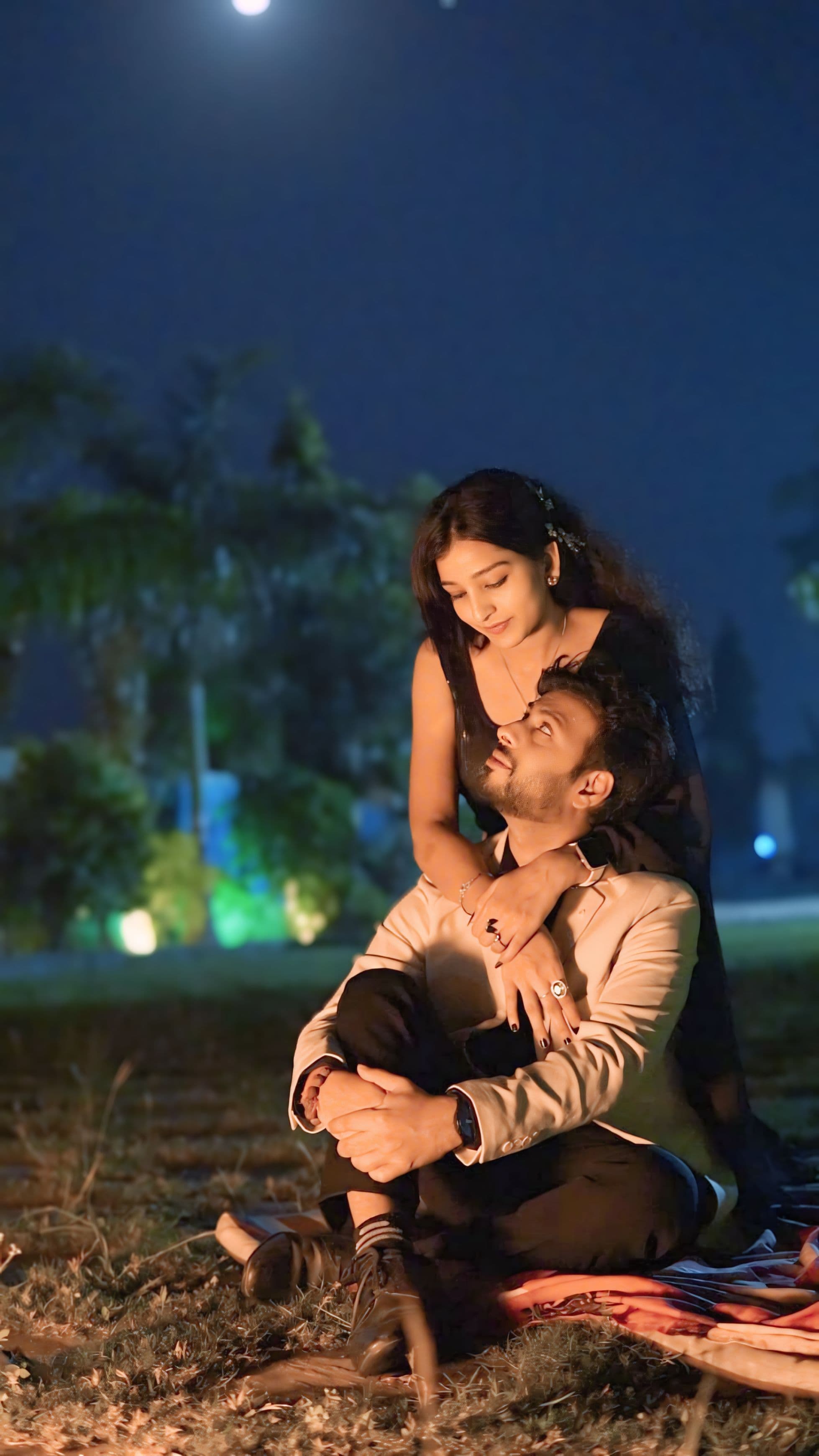 Couple sharing an intimate moment seated on the ground under the moonlight with soft bokeh lights in the background