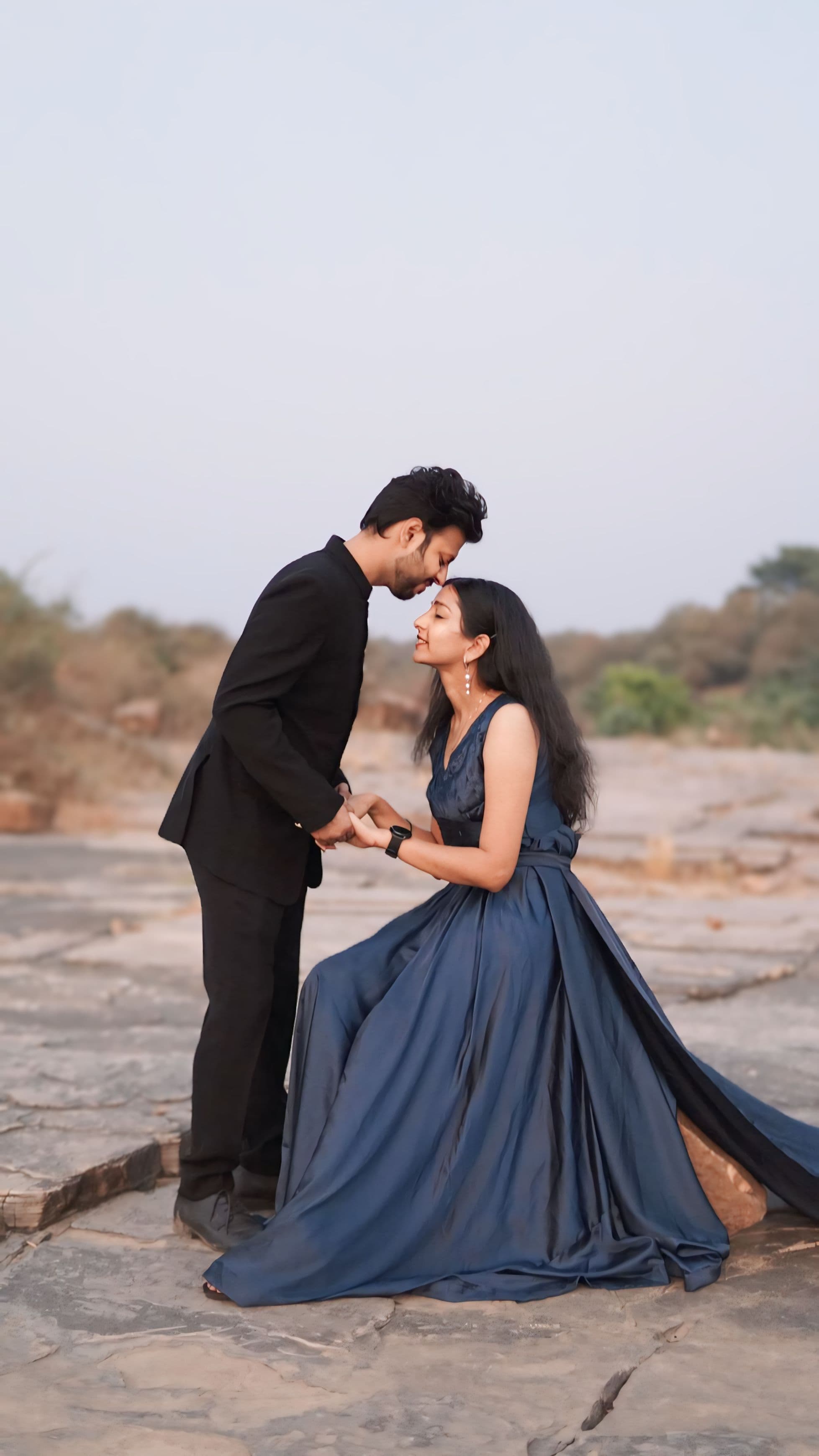 Elegantly dressed couple holding hands in a tender forehead touch on a rocky landscape at dusk