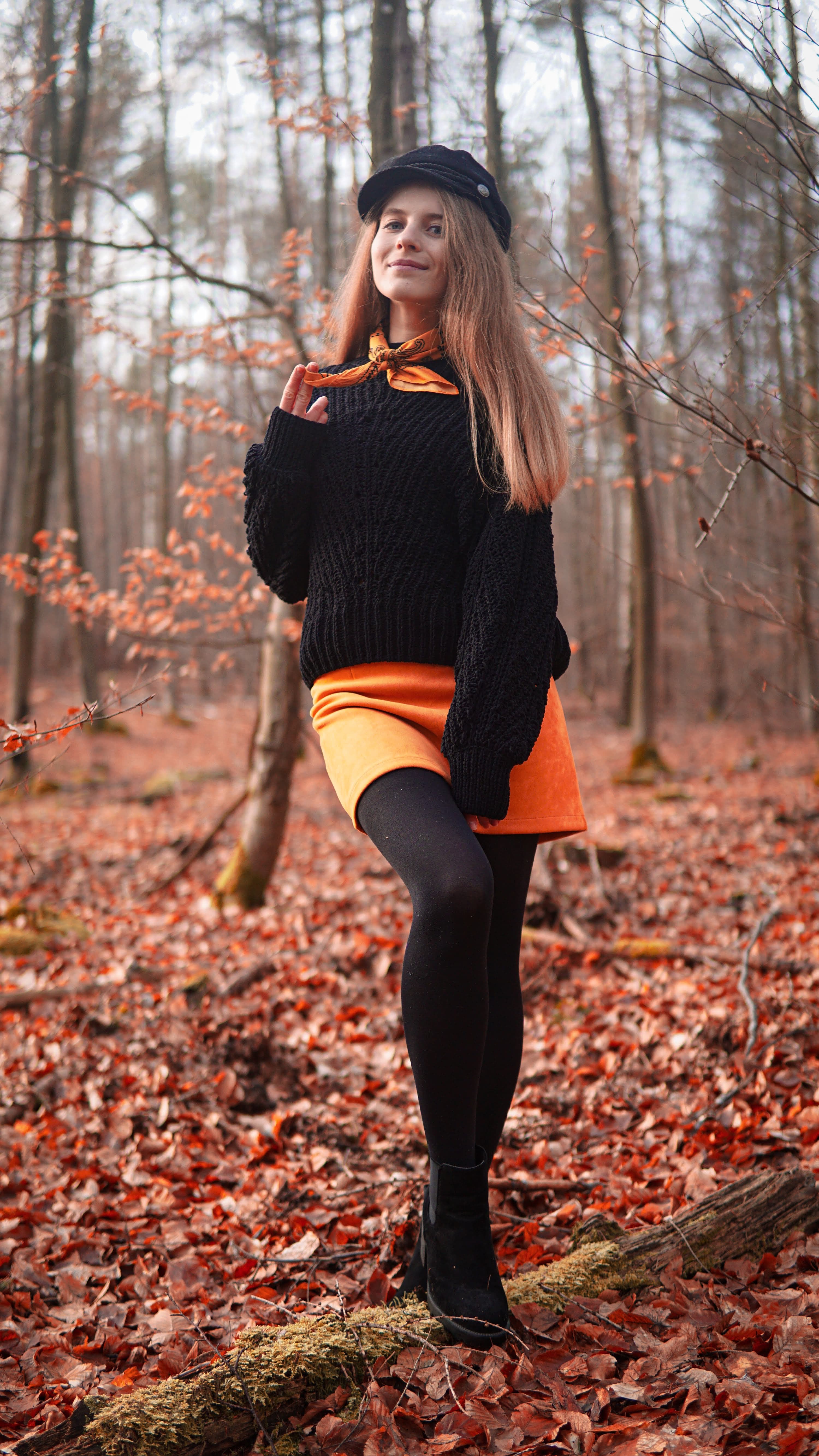 Full-length portrait of a woman in a black sweater and orange skirt standing on a log among fallen autumn leaves